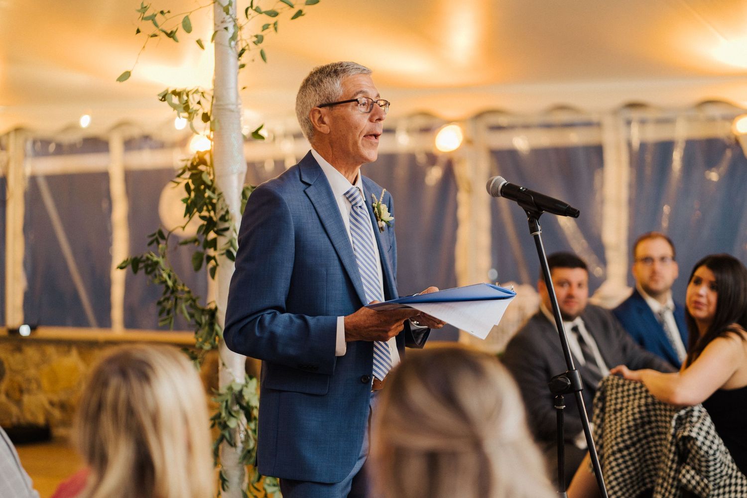 A speaker in a blue suit addresses wedding guests during an evening reception under a white tent.