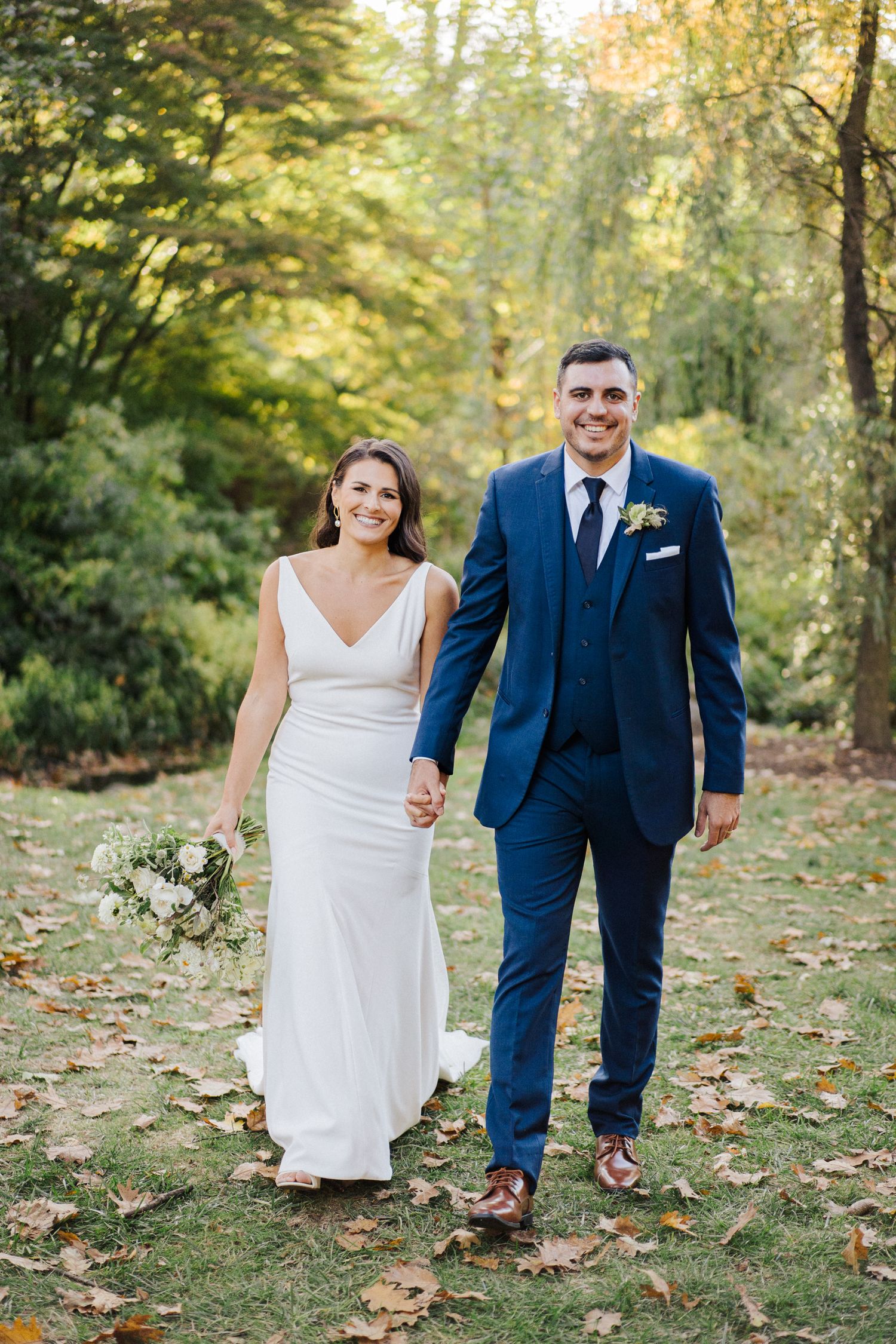 Newlyweds walk hand in hand through an autumn garden setting.