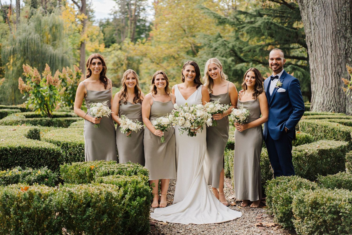 Wedding party poses in elegant garden setting with manicured hedges and fall colors.