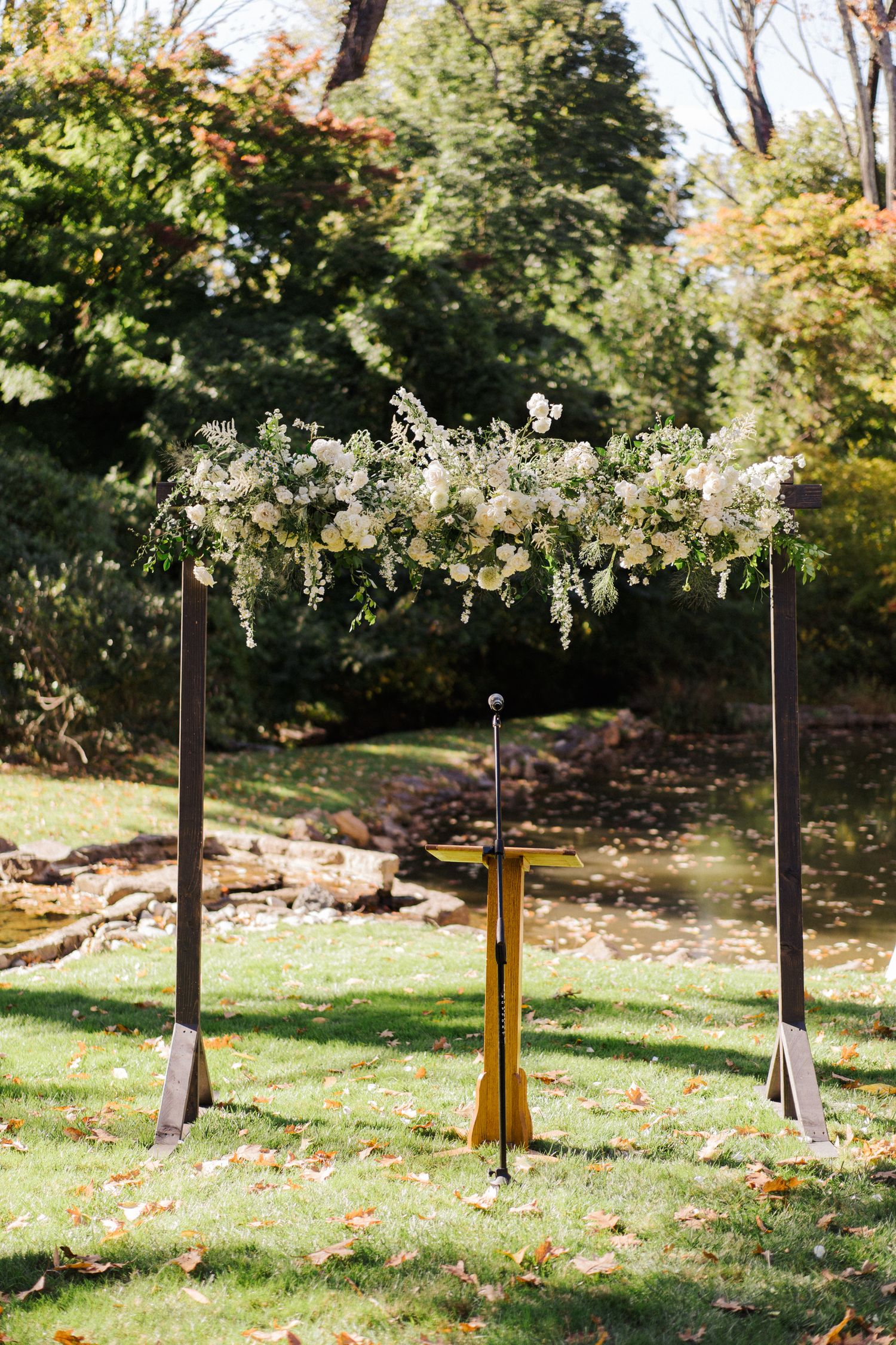Elegant floral wedding arch with white flowers stands on grass against autumn trees.
