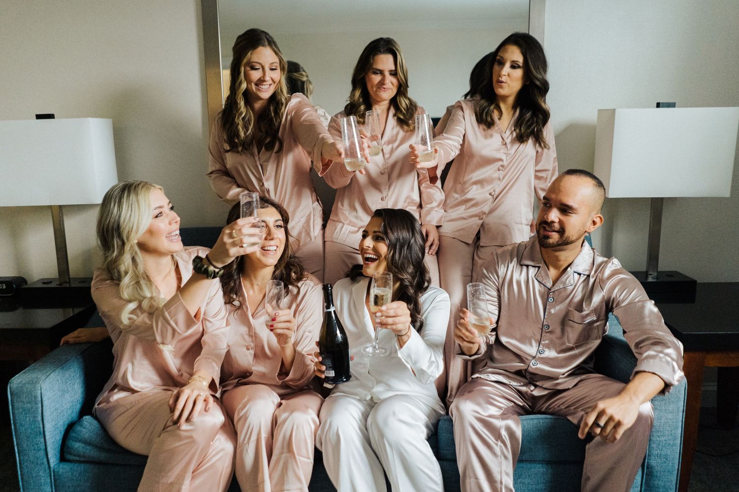 Wedding party relaxes together in matching pink silk pajamas on a bed with white pillows and lamps.