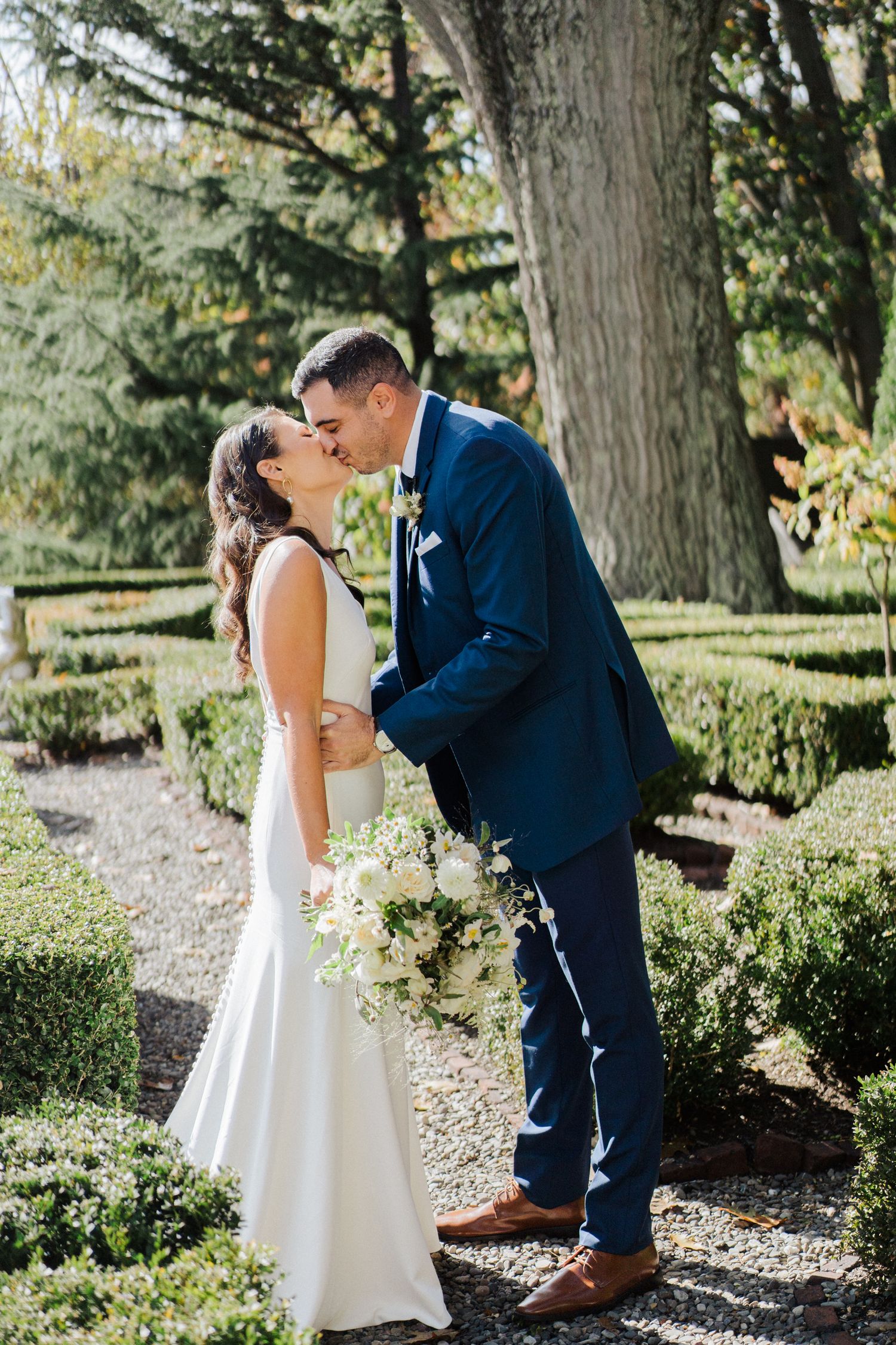 A tender wedding kiss photographed in a sunlit garden with beautifully landscaped surroundings.
