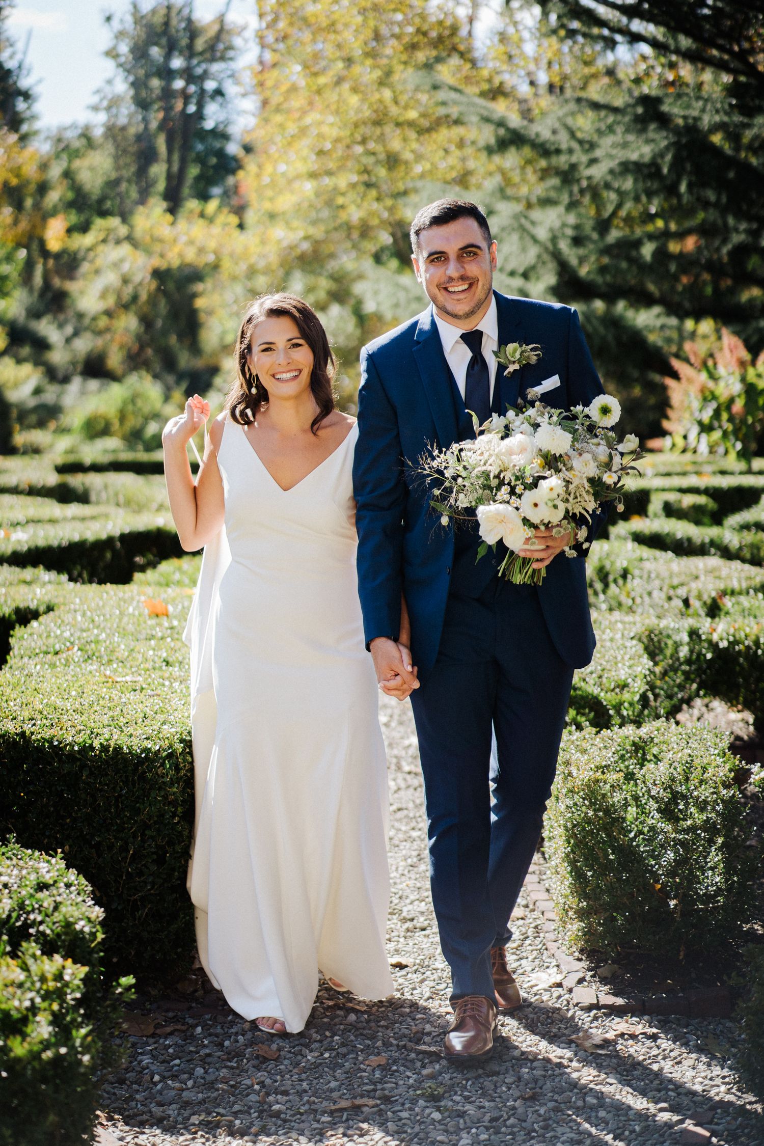 Wedding couple strolling hand in hand through garden pathway with white flower bouquet.