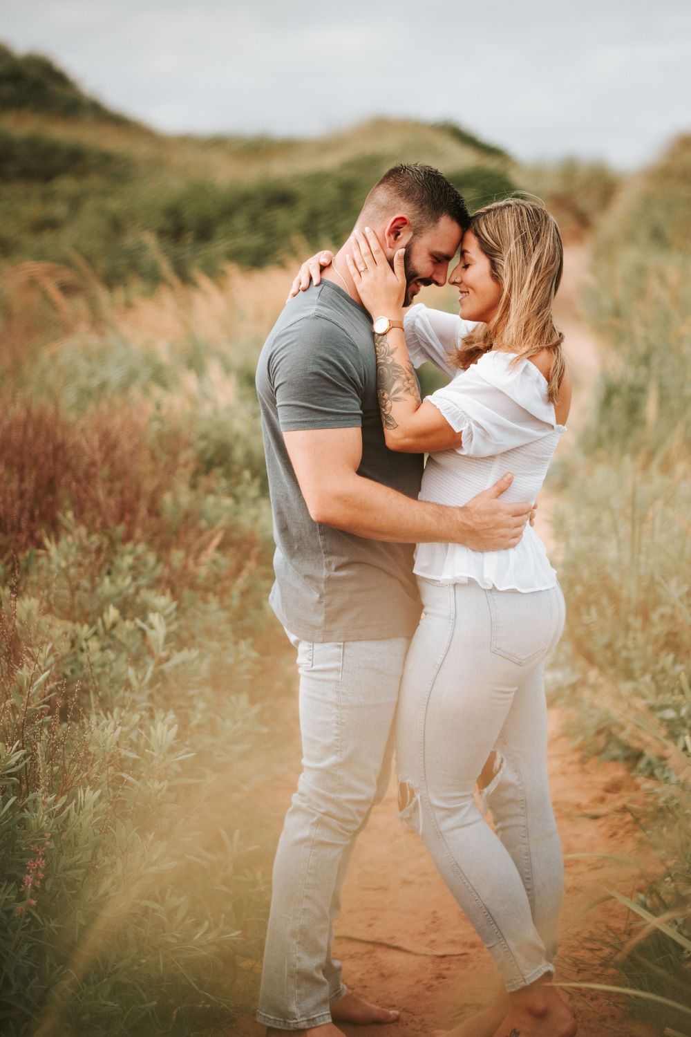 A couple shares an intimate moment on a dirt path surrounded by coastal vegetation.
