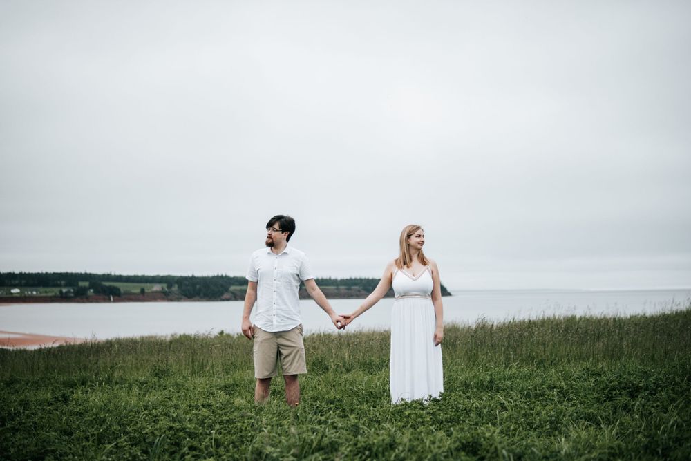 A couple in white clothing holds hands while standing on a grassy clifftop overlooking the ocean.