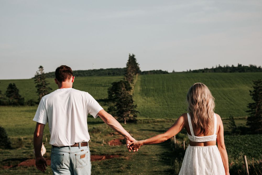 A couple holds hands while walking through a green summer field with ocean views.