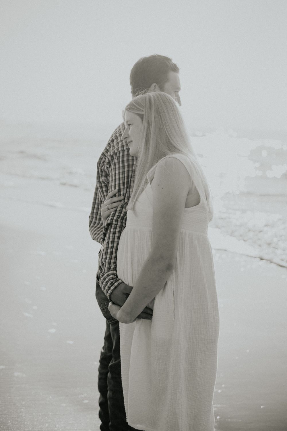 A couple stands close together on a misty beach in black and white.