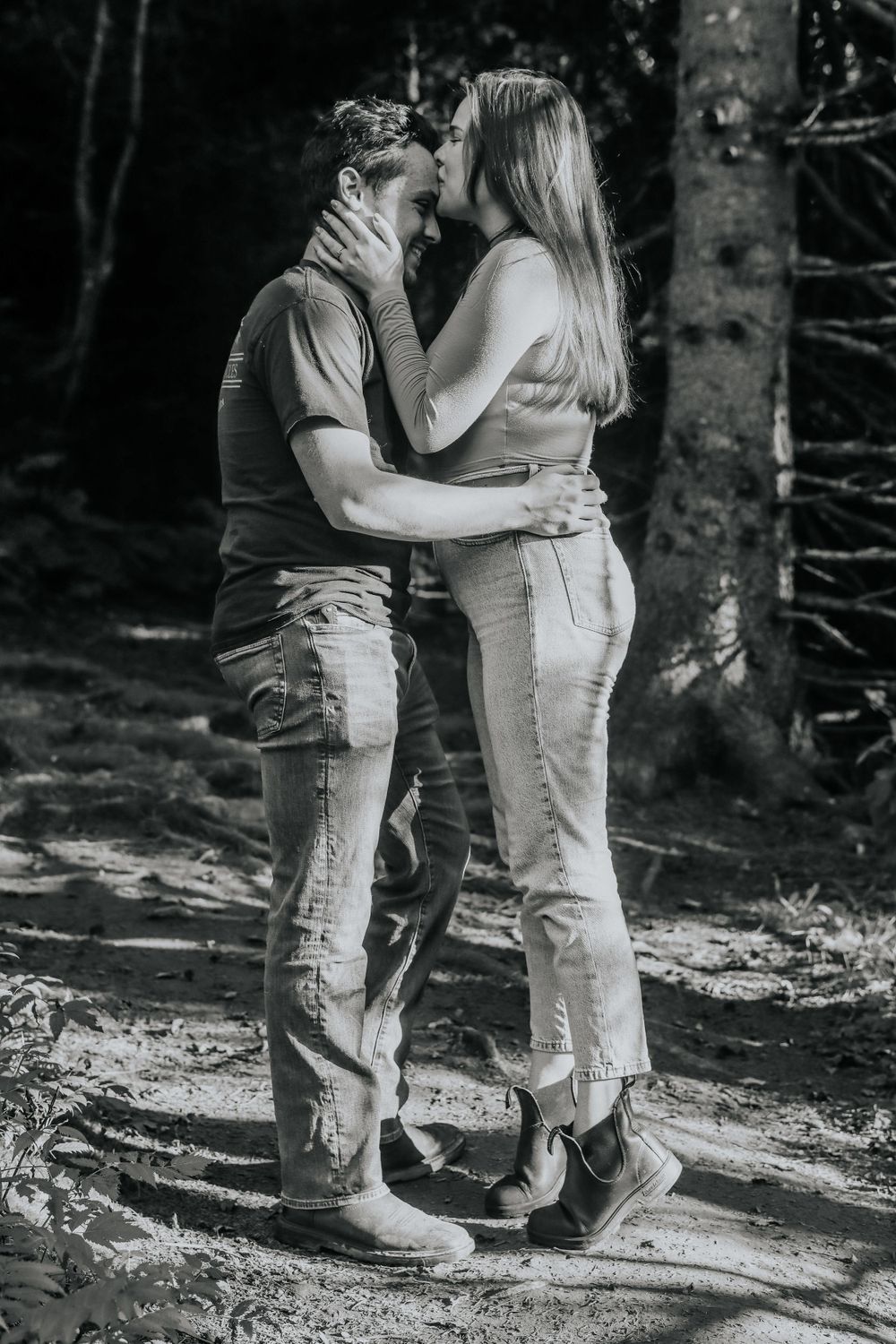 A couple shares an intimate moment on a forest path in black and white.