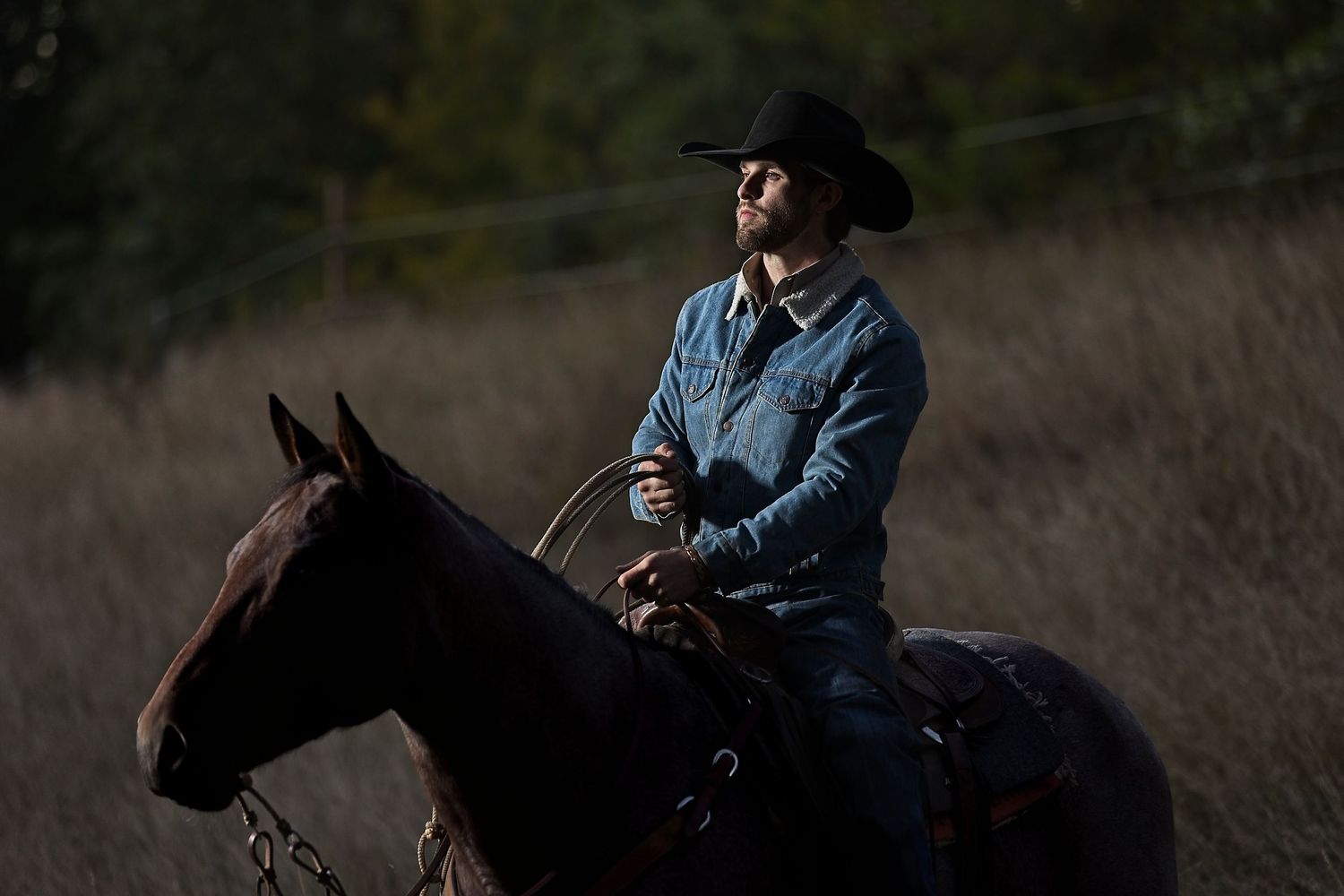 A rider in a denim jacket and black cowboy hat sits atop a dark horse in moody evening light.