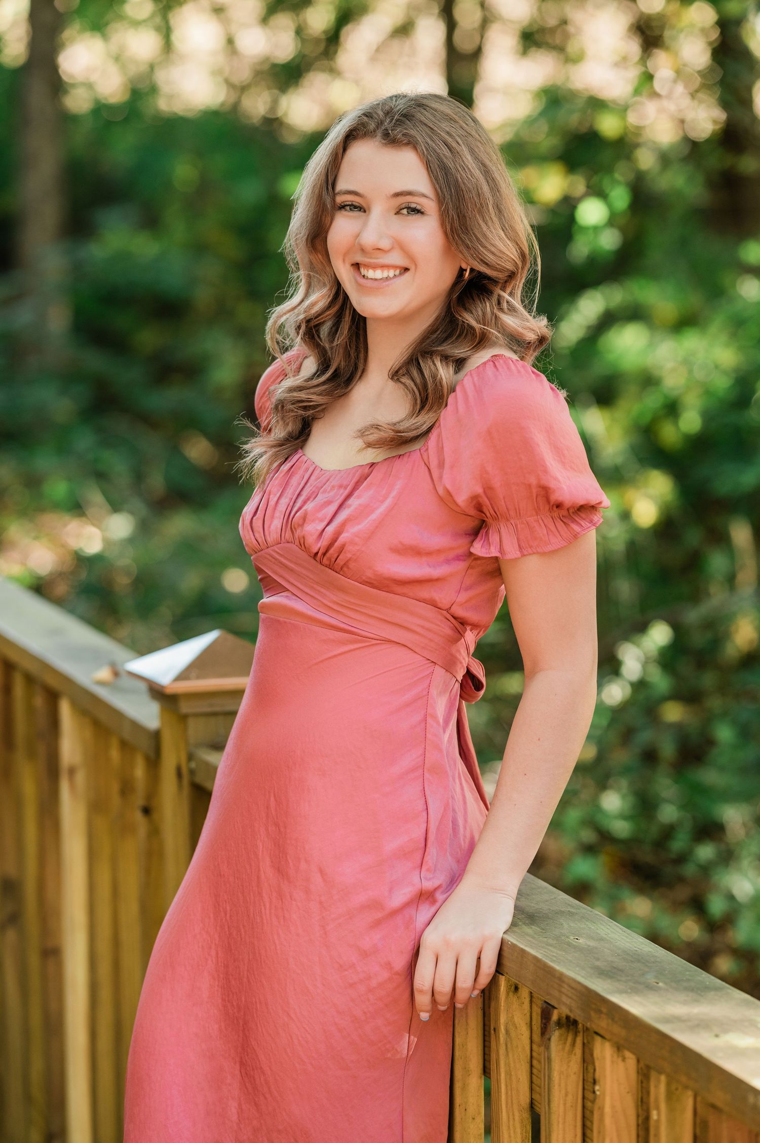 Outdoor portrait series in coral dress leaning against wooden railing with natural greenery backdrop.