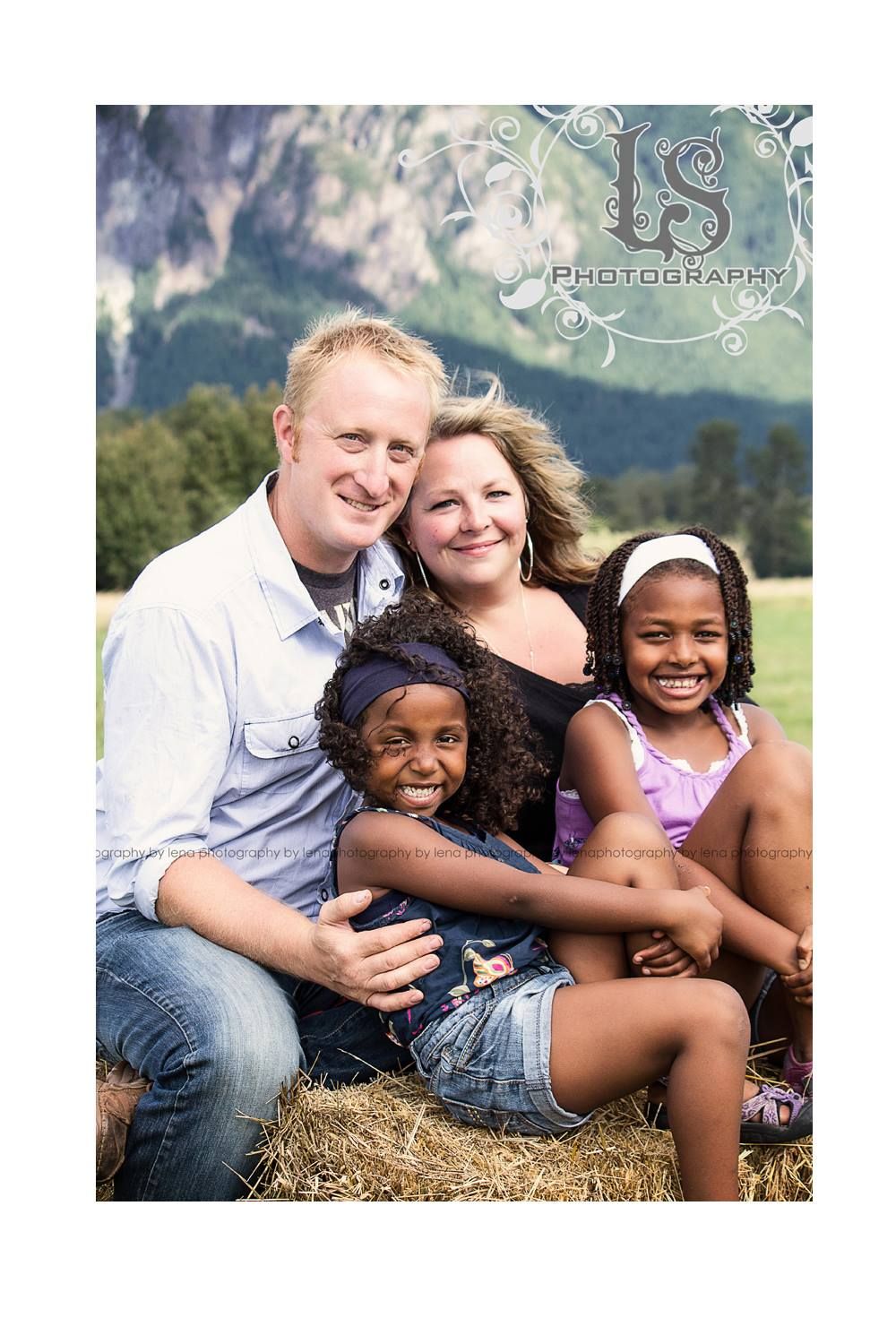 Family relaxing on hay bale with mountain backdrop during outdoor photo session.