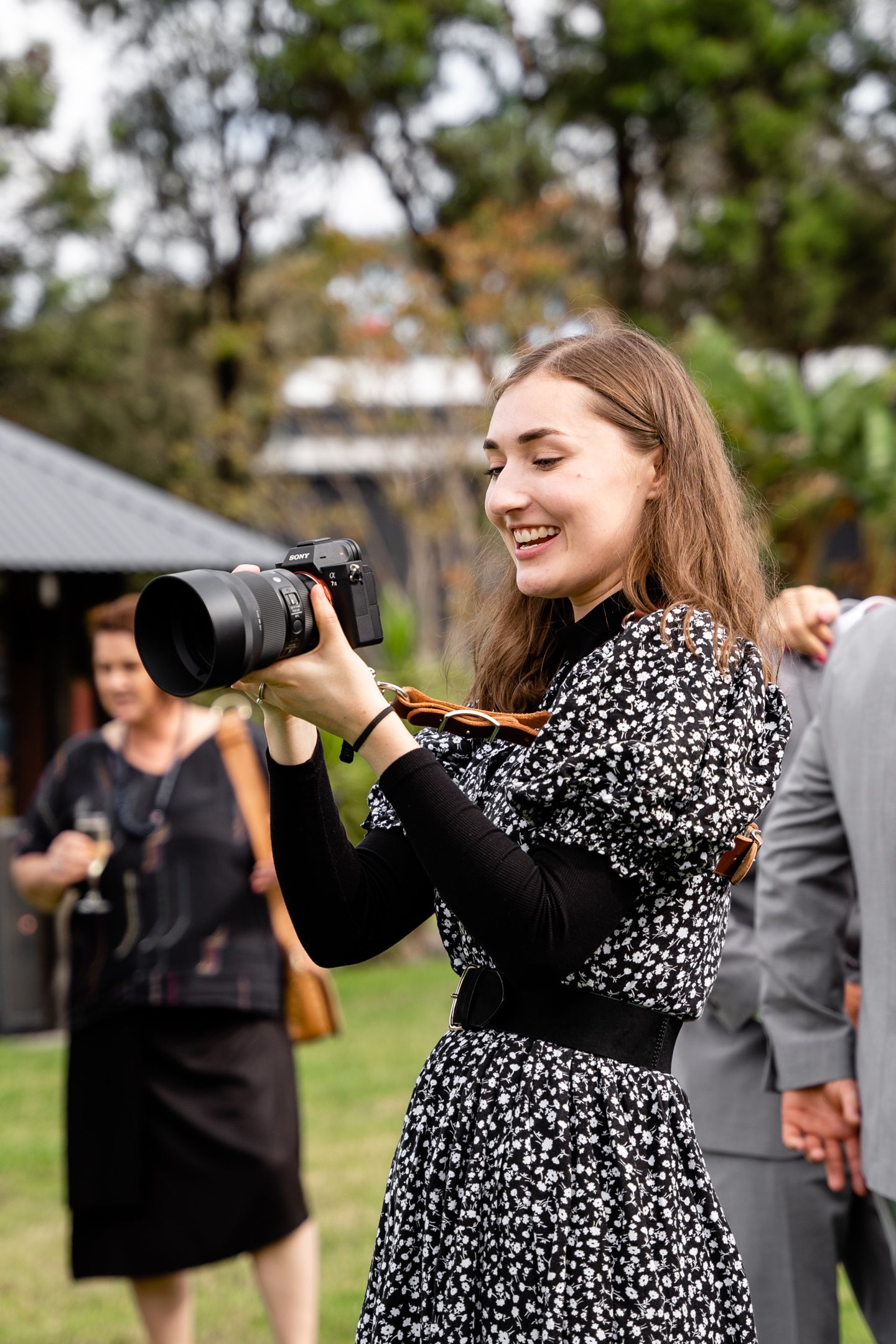 Photographer in a floral dress capturing photos at an outdoor event with greenery in background.