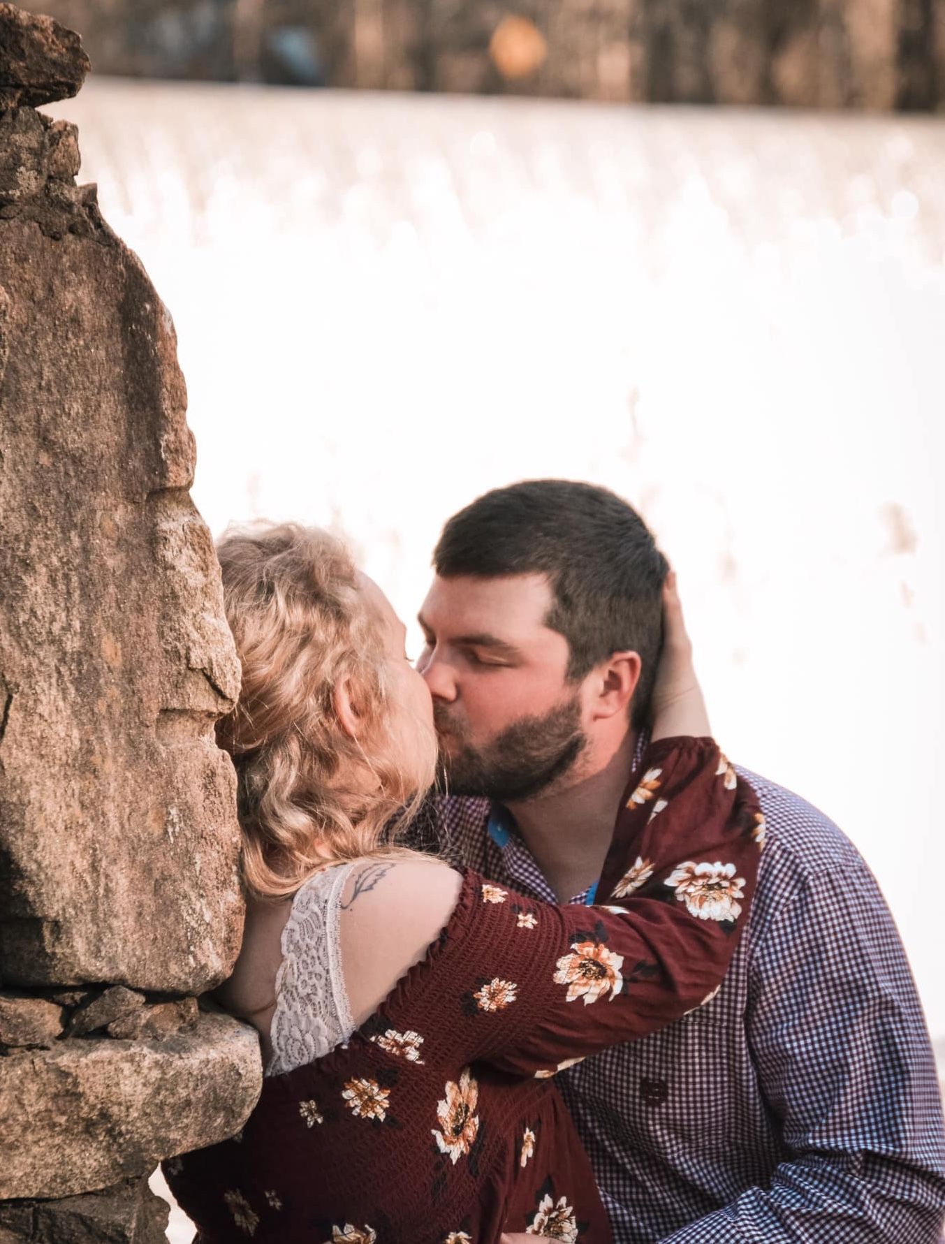 Couple shares intimate moments in a rustic outdoor setting with stone wall backdrop.