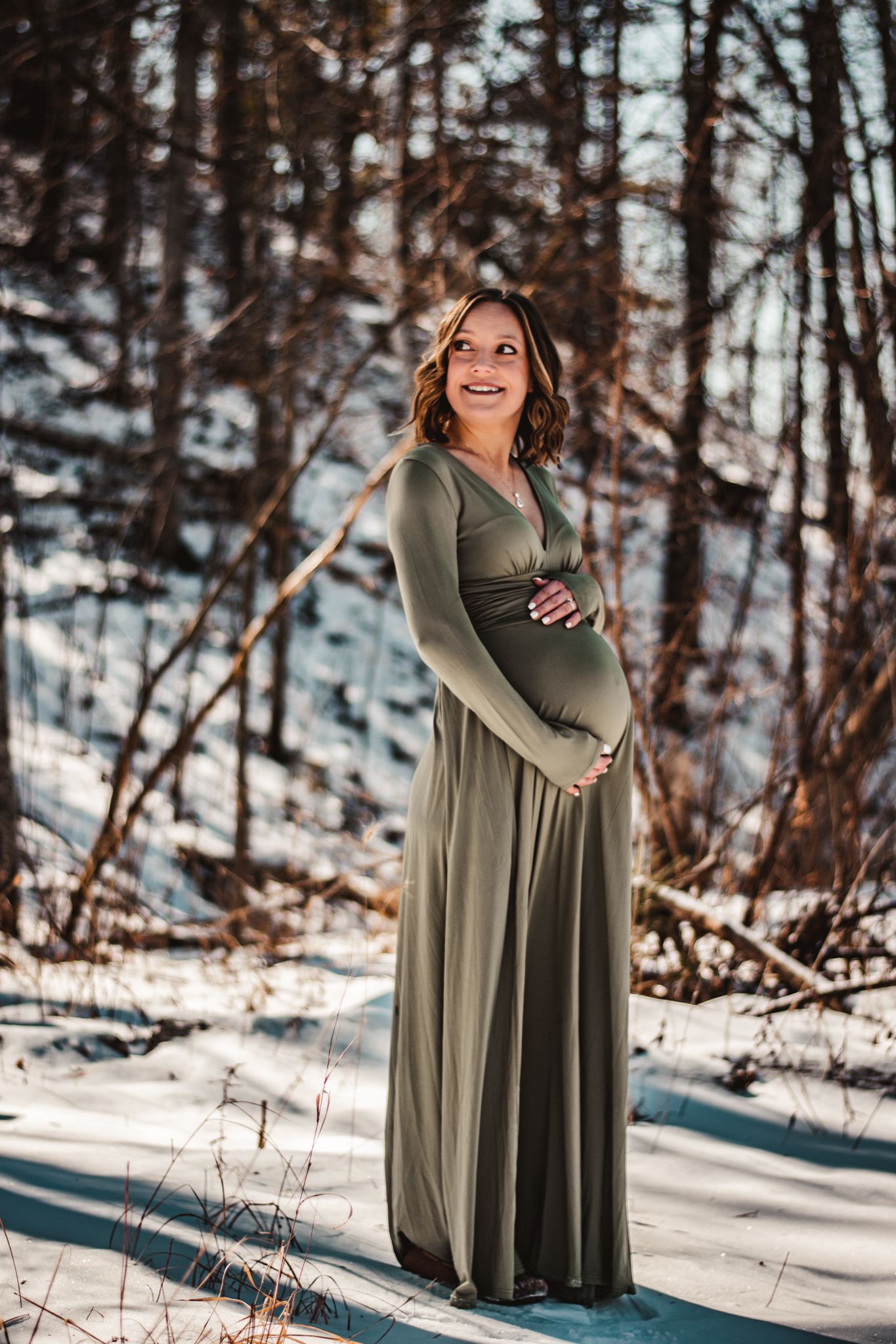 Winter maternity photo of a woman in a olive green dress standing in snowy woods.