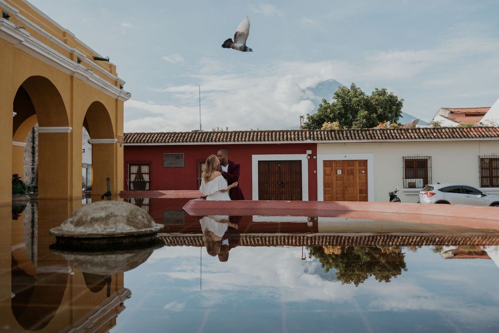 A reflection shot of a couple by a colonial building with arches and water.
