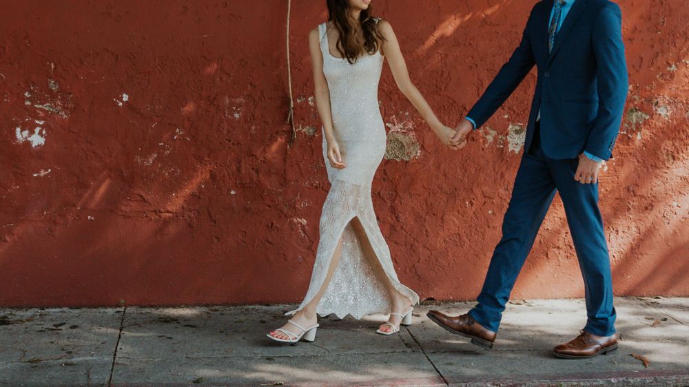 A couple in formal attire walks hand in hand along a red wall.