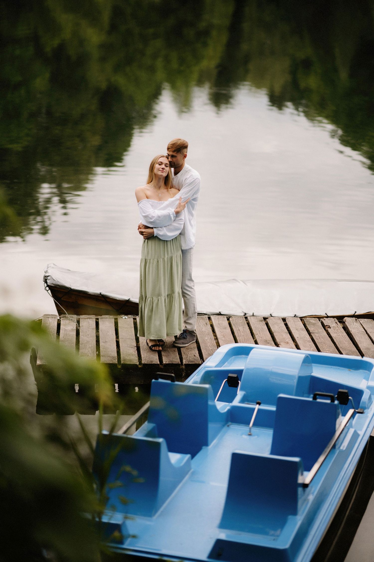A couple embracing on a wooden dock by a lake with a blue pedal boat in view.