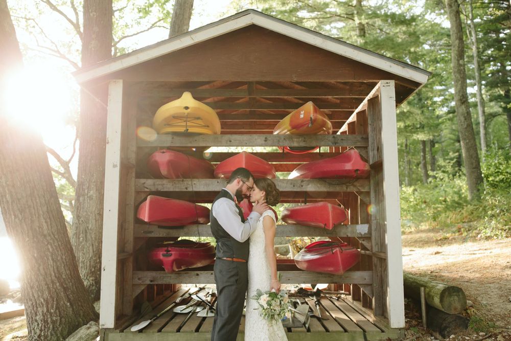A couple embraces in beautiful natural light at their  NH summer camp wedding. A rustic shed with canoes and kayaks in the background.