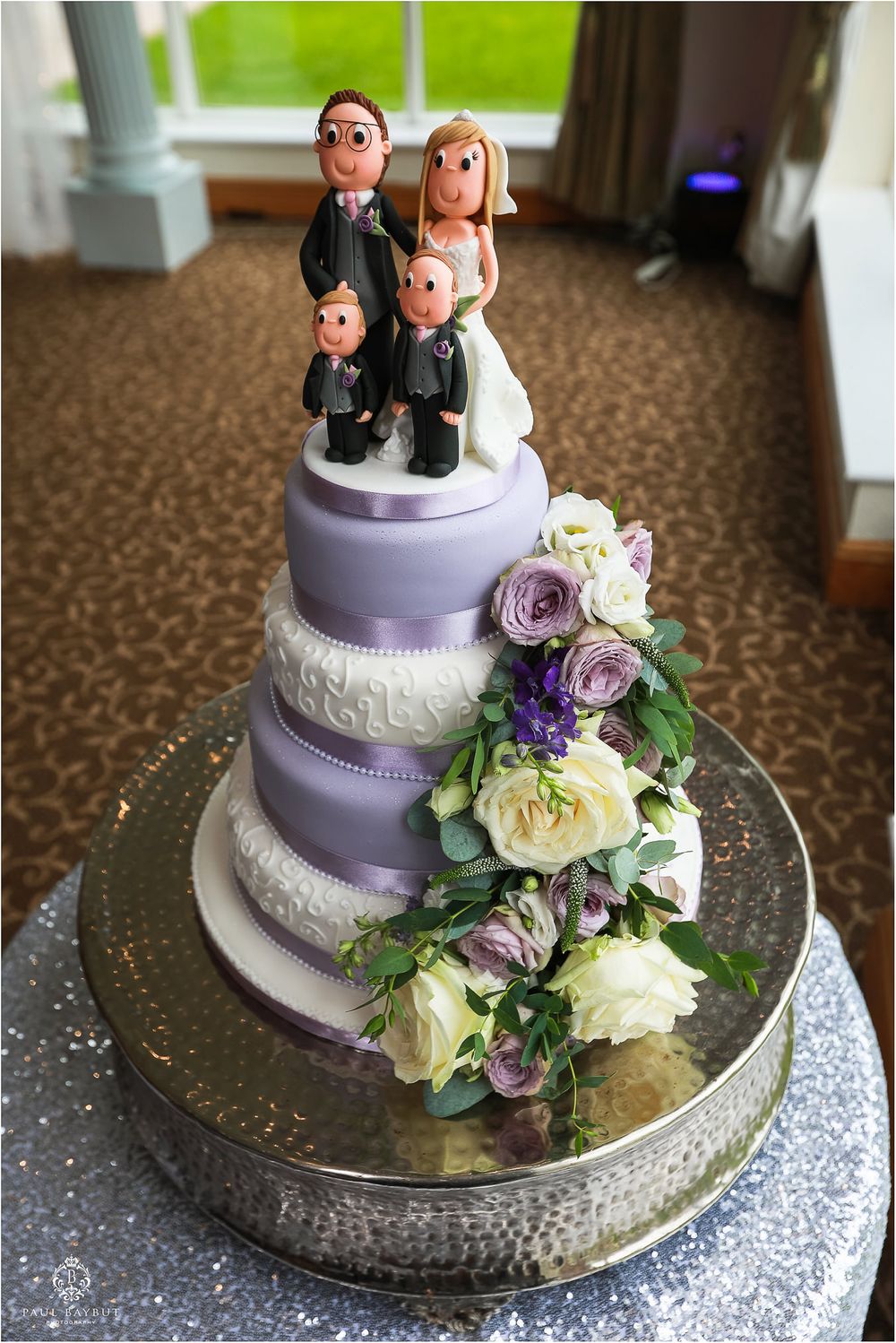 Purple and white tiered wedding cake with custom family topper and fresh floral decorations.