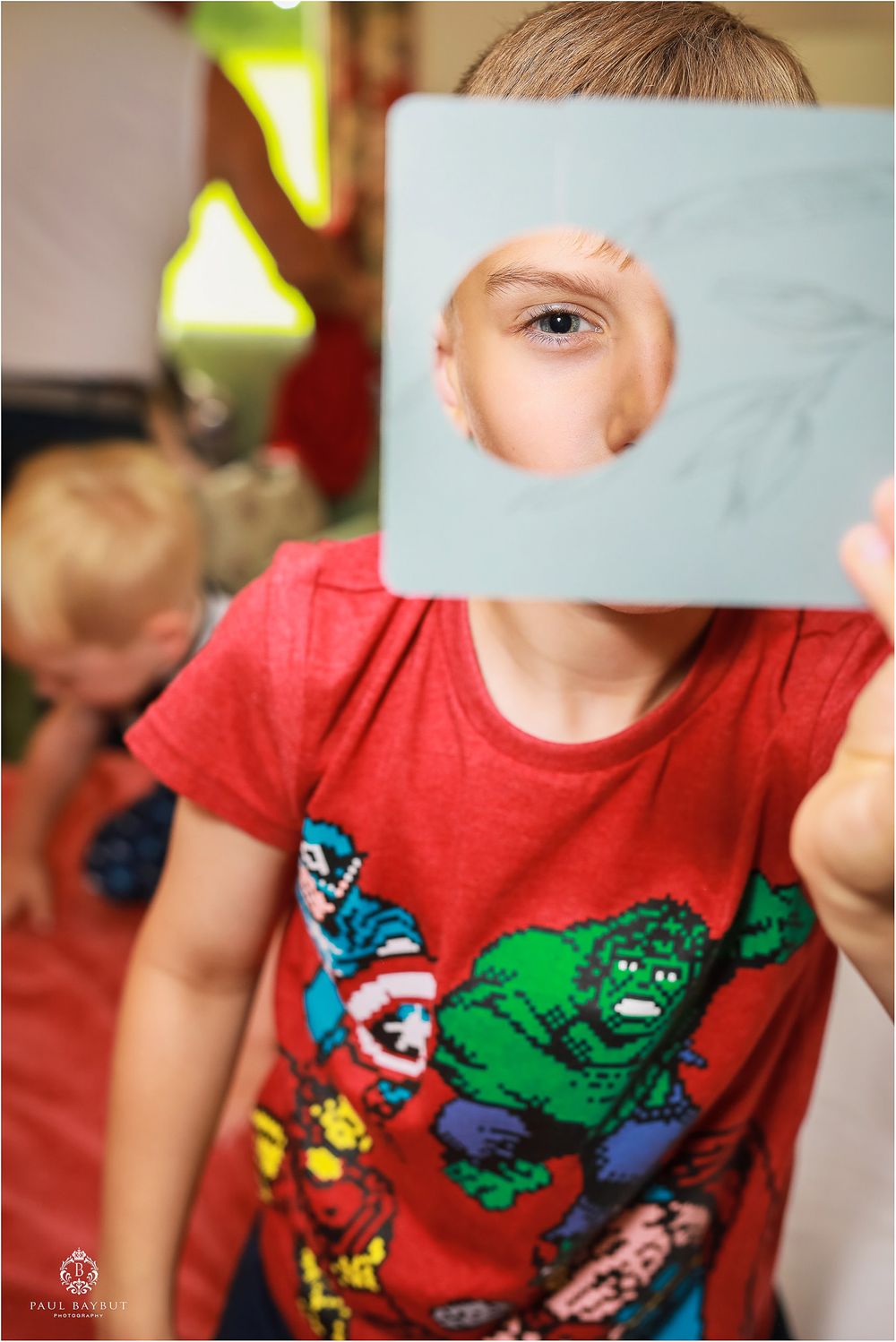 A child in a superhero shirt looks through a square cutout in blue paper.