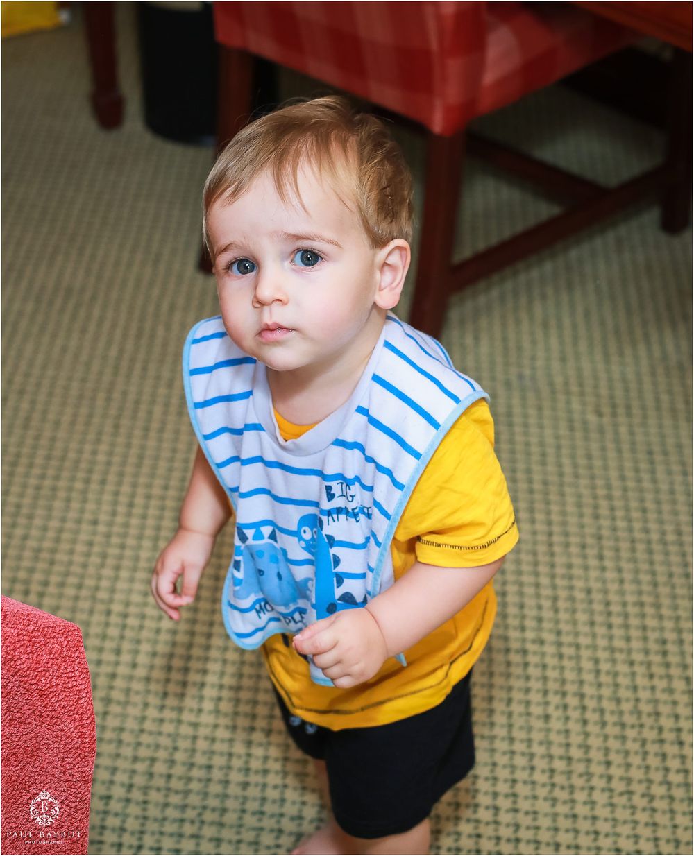 A toddler wearing a striped bib and yellow shirt stands on carpet.