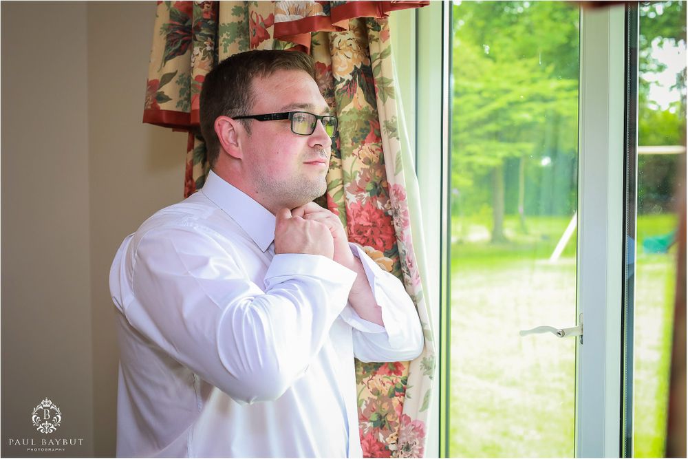 A man in white shirt and glasses adjusts his collar by a window.