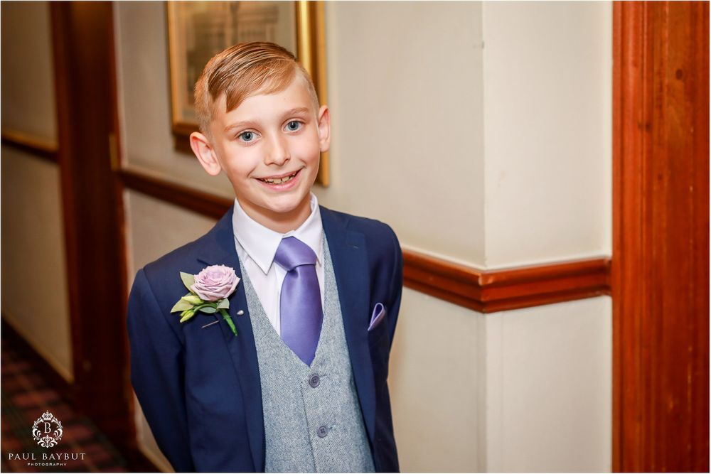 A young boy wearing a navy suit with pink boutonniere smiles in a hallway.