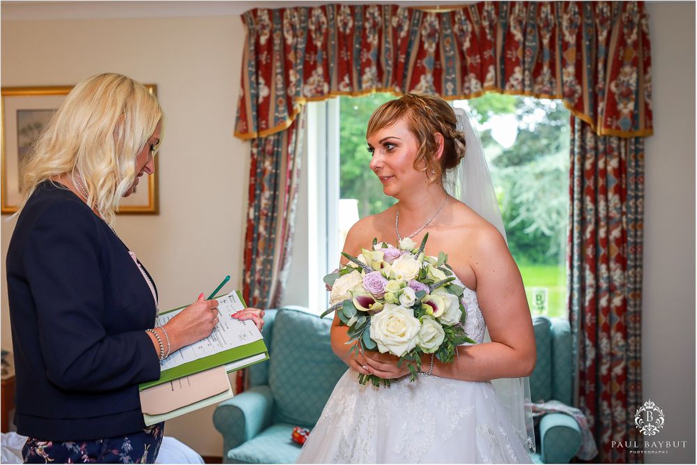 Bride holding bouquet speaks with wedding coordinator in decorated room.