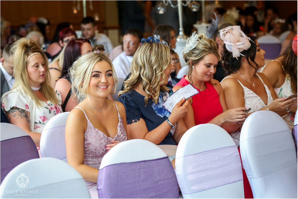 Wedding guests seated in white-covered chairs enjoy the ceremony in an elegant reception hall.