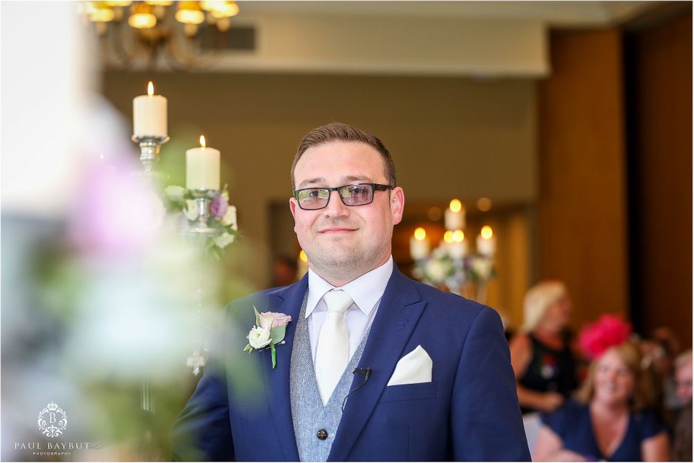 Groom in navy suit with white tie and boutonniere stands before ceremony backdrop with candles.