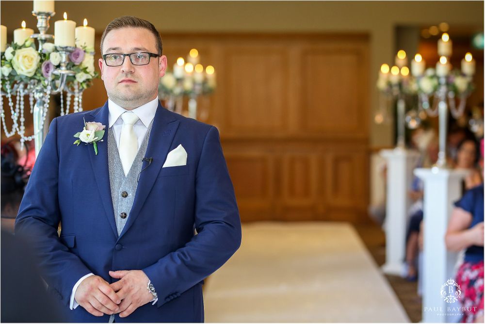Groom in classic navy suit awaits at altar with elegant candlelit ceremony decor.