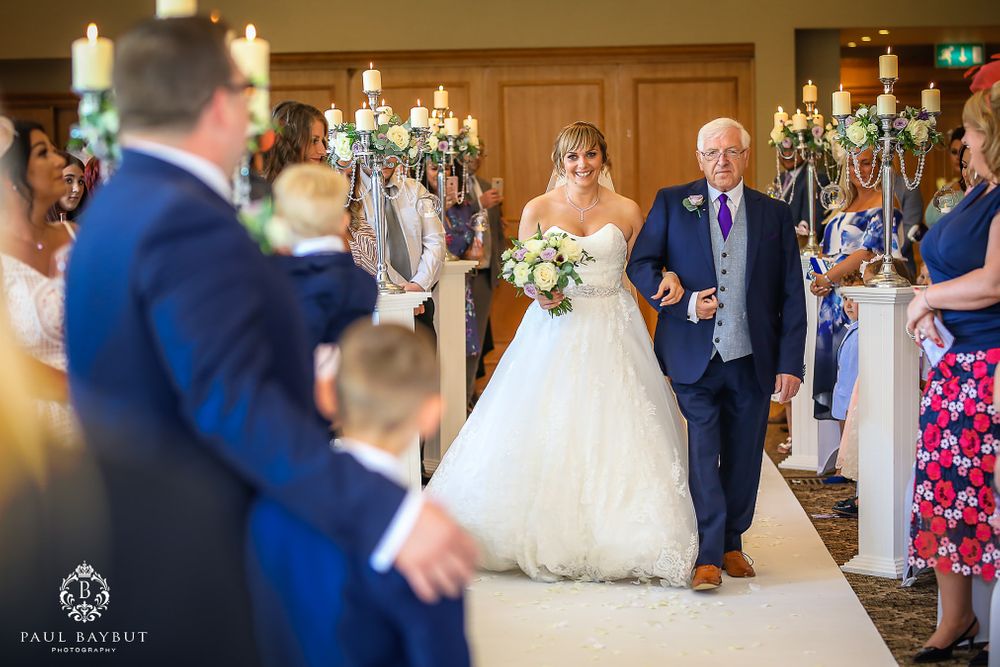 Wedding ceremony procession in elegant hall with white candelabras and purple decor accents.