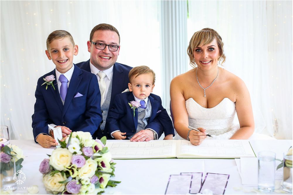 Wedding party signing official documents with floral arrangements on white table.