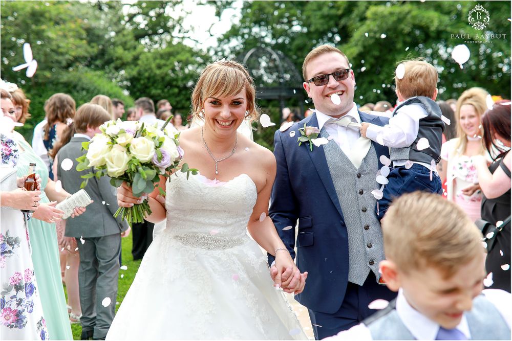 Newlyweds walking outside with classic white bouquet among wedding guests.