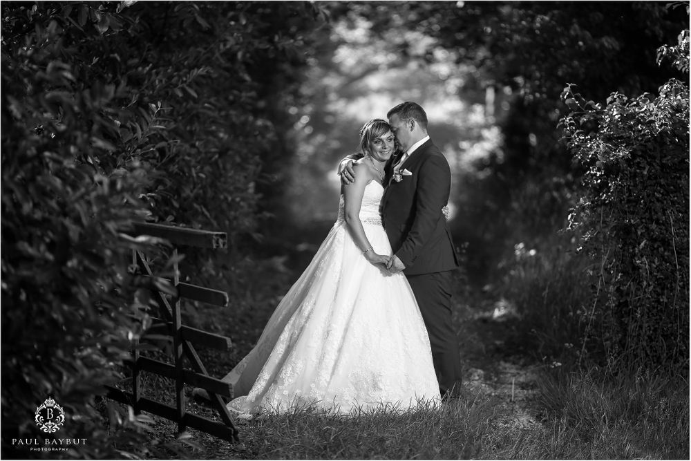 Wedding couple shares a tender moment beside a wooden bench in a black and white photograph.