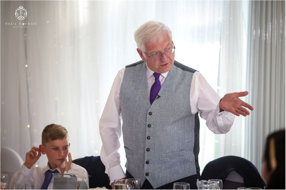 Speaker wearing a grey vest and purple tie addresses guests at a wedding reception.
