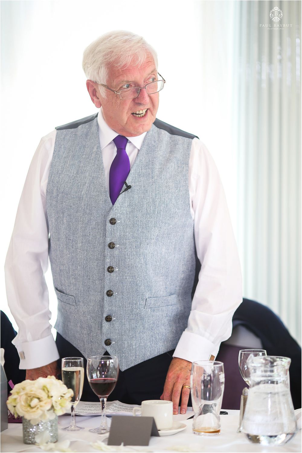 Speaker in grey vest and purple tie smiles while addressing wedding guests.