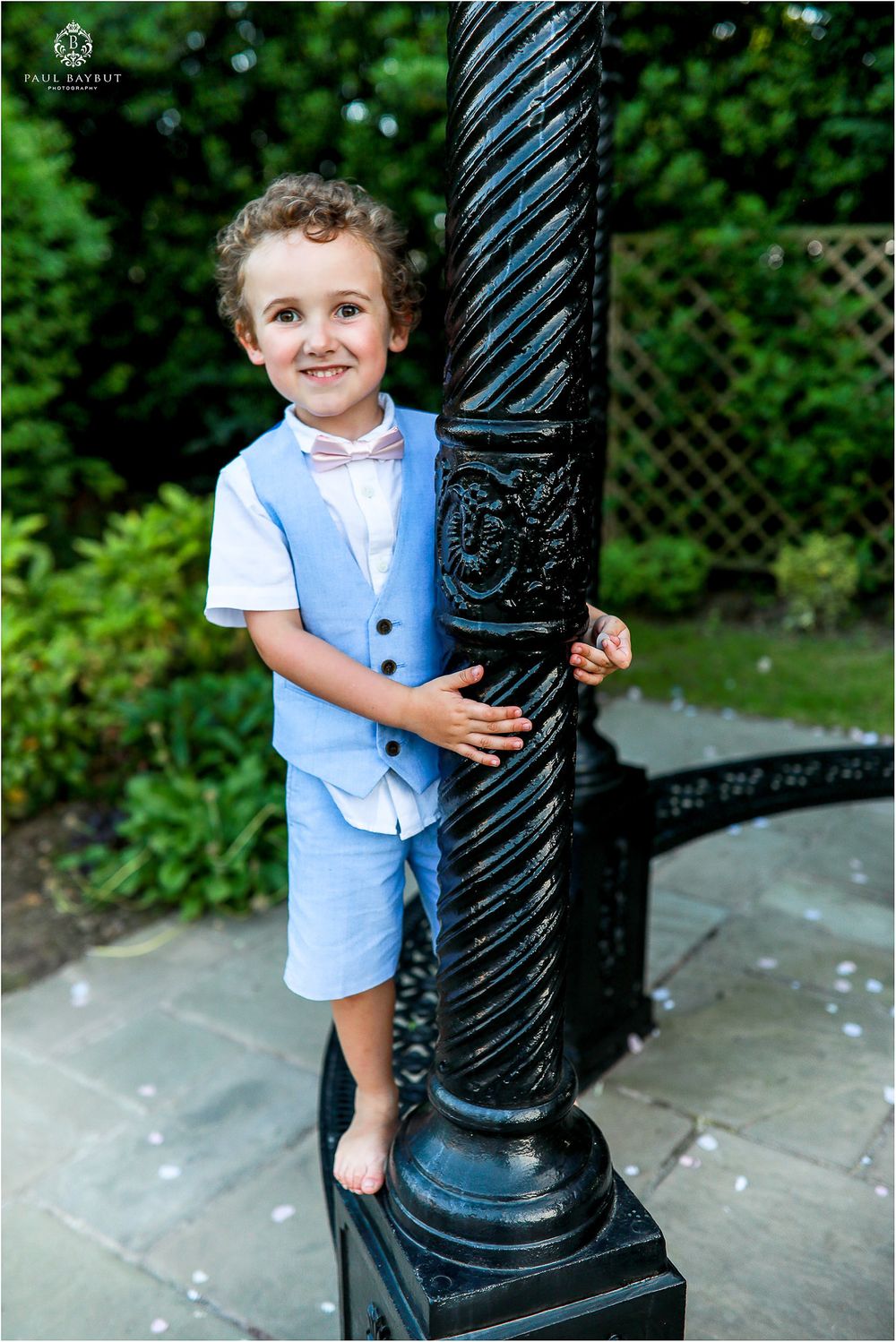 Young person in a light blue outfit poses by a decorative black metal pole in a garden.