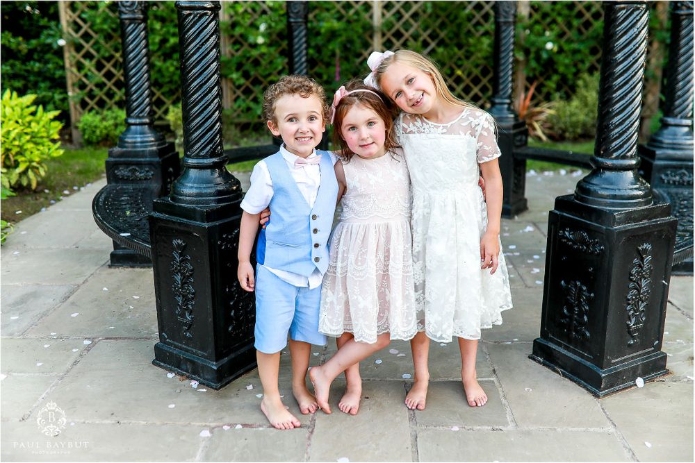 Three children in formal attire pose between ornate black posts outdoors.