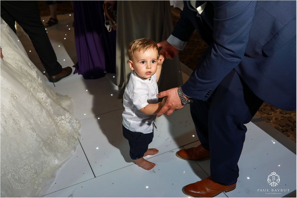 Small toddler in formal wear holds hands with an adult on a white dance floor at a wedding reception.