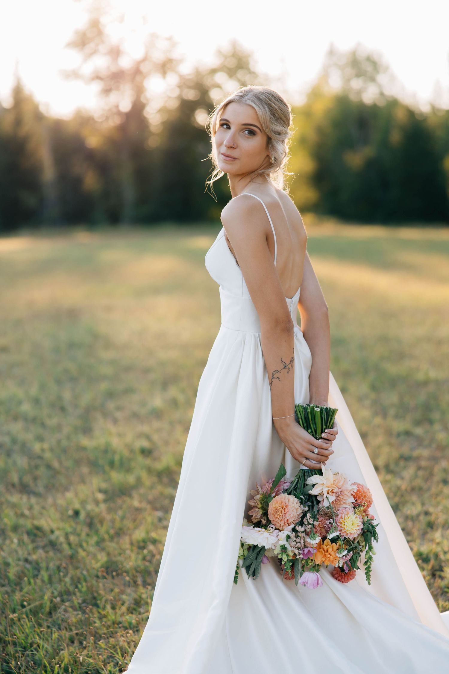 Elegant bride in backless white gown holding a colorful bouquet in a sunlit green meadow at sunset.