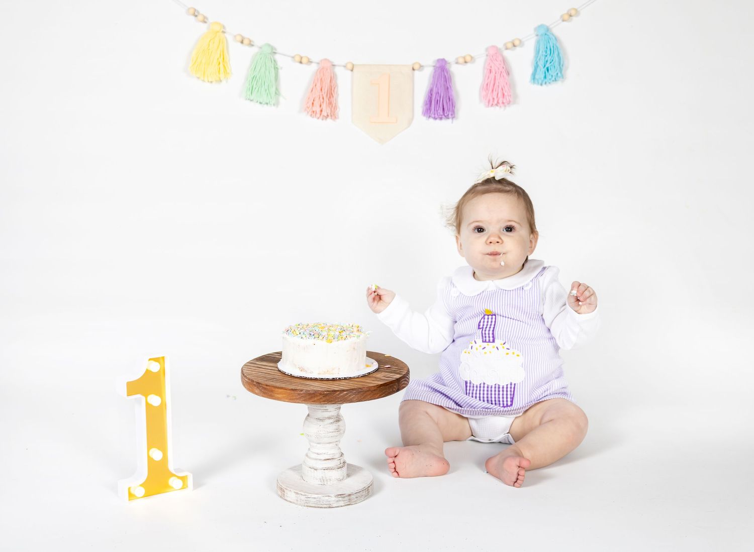 A smiling baby sits on a white backdrop with colorful party decorations and a cake for their first birthday cake smash.