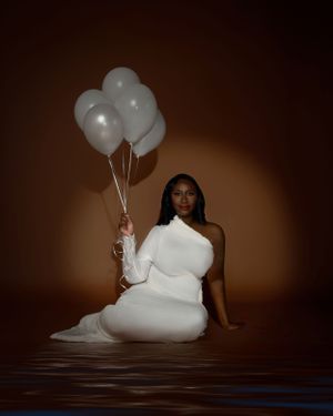 A woman in a white dress sits elegantly with silver balloons against a dark brown backdrop and spotlight for a birthday photoshoot.