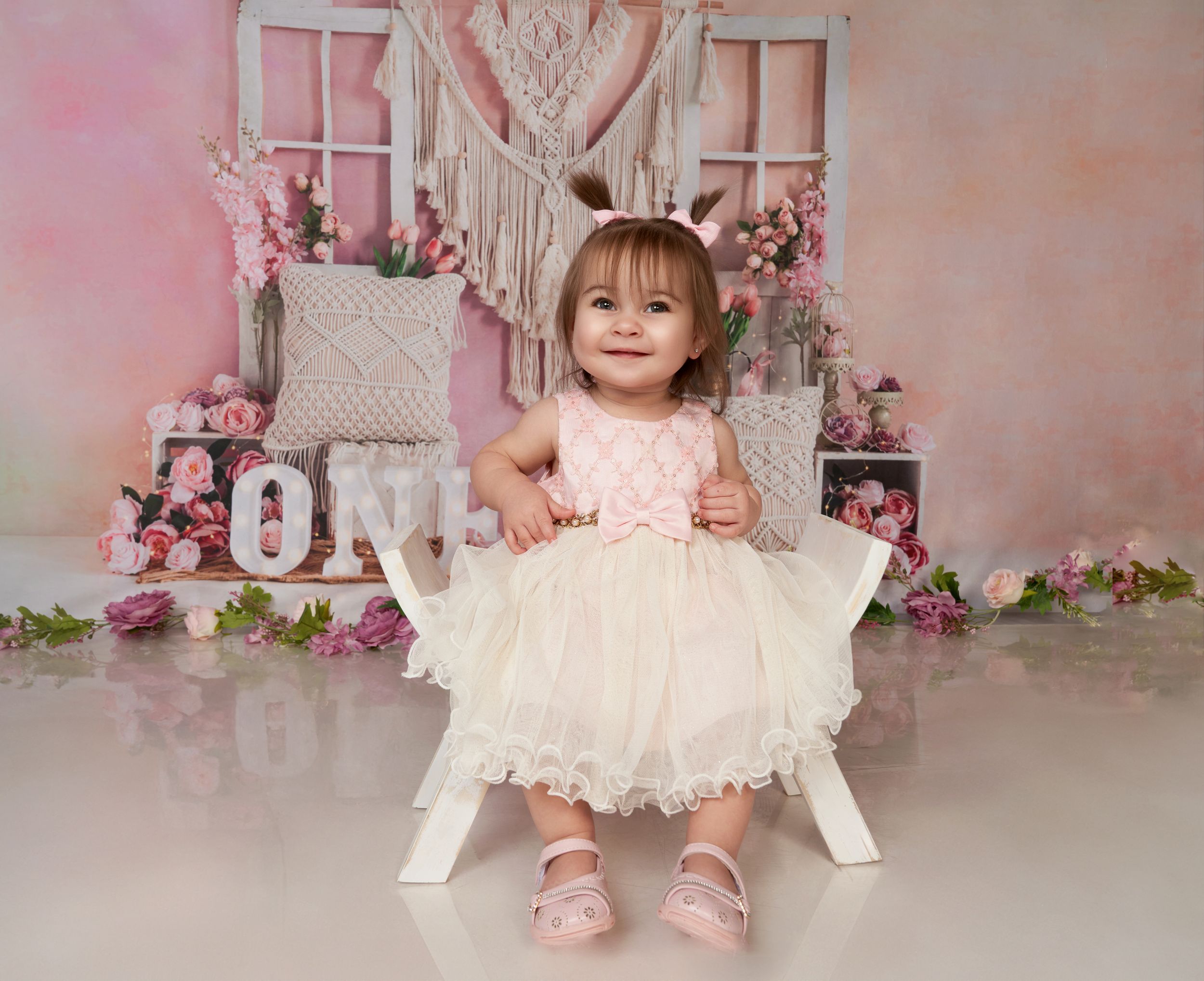Elegant first birthday photo setup with white chair, pink floral decorations, and lace dress against soft pink backdrop.
