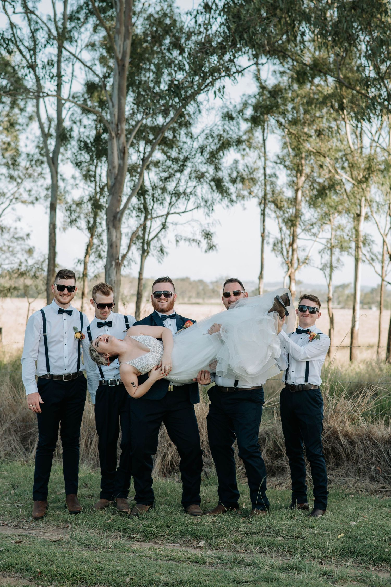 Wedding party members in suspenders and sunglasses hold someone up in a fun group pose.