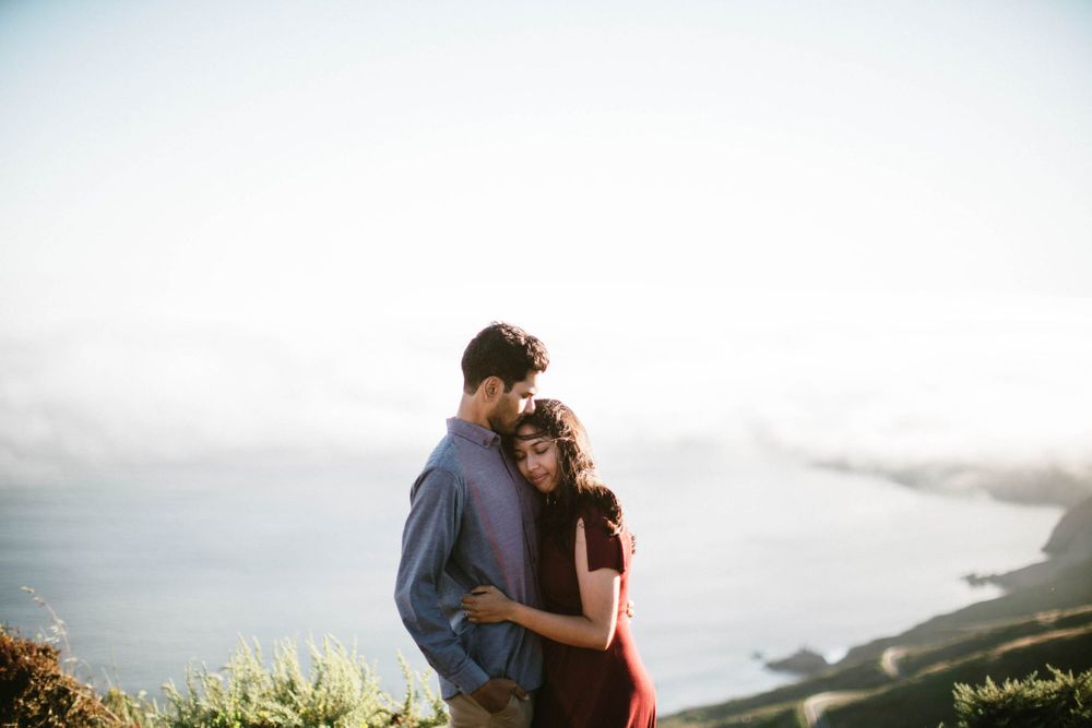 Couple embracing on coastal hilltop with dramatic fog rolling over ocean below.