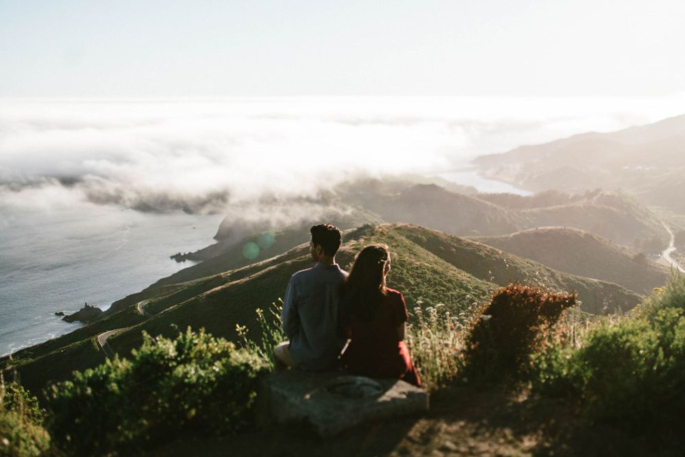Pair sitting together on mountain cliff edge overlooking dramatic coastal fog.