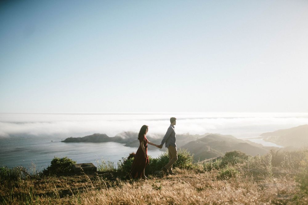 Couple stands atop scenic mountain overlooking foggy coastal landscape at sunset.