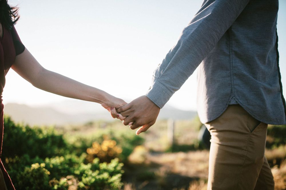 Close-up of two people holding hands against natural outdoor backdrop.
