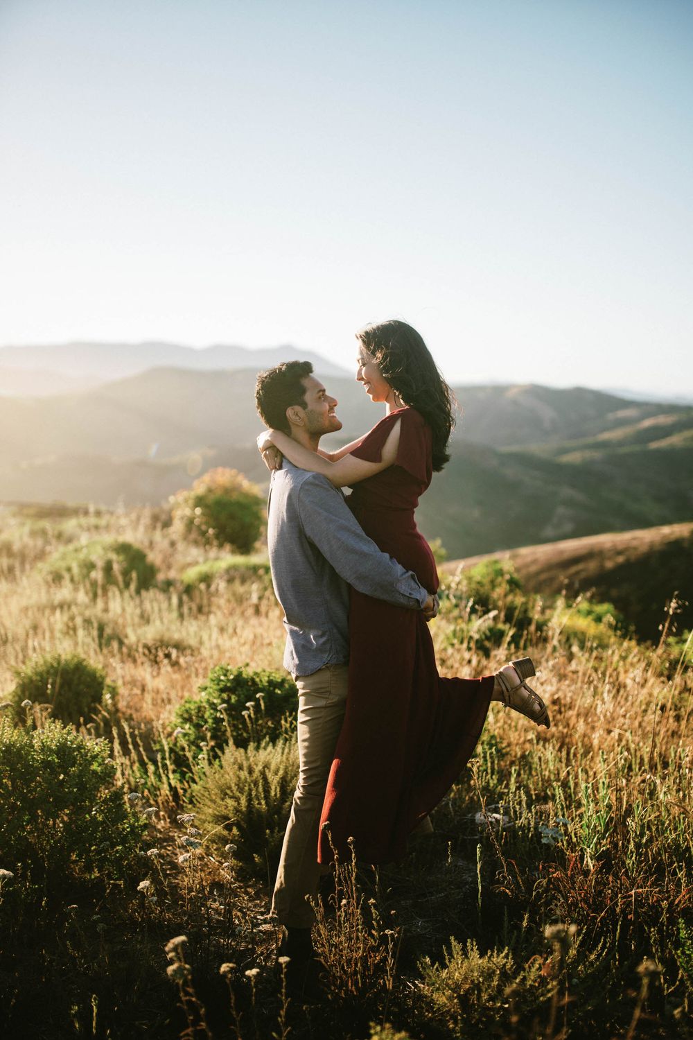 A romantic couple embraces on a hilltop at sunset with mountains in the background.